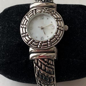 May include: Silver-toned bangle watch with a round face and a white mother-of-pearl dial. The watch features a textured bezel and band, with a decorative top piece. The watch has a classic, elegant design.