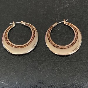 May include: A pair of gold-toned hoop earrings. Each earring has a wide, polished outer band and a narrower inner band filled with small, brown beads. The earrings are displayed on a dark, textured surface.