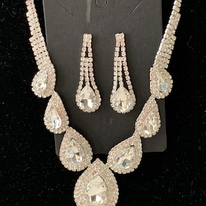 May include: A silver-toned jewelry set featuring a necklace and matching earrings. The necklace and earrings are adorned with clear, teardrop-shaped rhinestones. The necklace has a series of larger teardrop stones and smaller stones along the chain. The earrings are a dangling design.