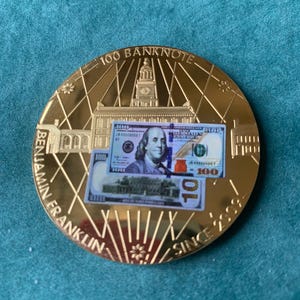 May include: A gold-colored commemorative coin featuring a depiction of a $100 bill and Benjamin Franklin. The coin has the text "100 BANKNOTE" and "BENJAMIN FRANKLIN SINCE 2008" around the edge.