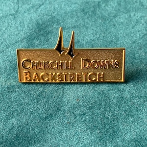 May include: A gold-colored pin with the text "CHURCHILL DOWNS BACKSTRETCH" in capital letters. The pin features a stylized design of two black and gold horse racing posts. The pin is rectangular in shape.