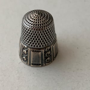May include: A silver thimble with a textured, perforated dome and a decorative band at the base. The thimble's design features a series of small holes for needlework and ornate scrollwork on the band. This sewing tool is designed to protect the finger while sewing.