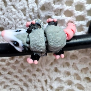 Puede incluir: Un pequeño amuleto de zarigüeya gris y blanco con detalles rosas. El amuleto está unido a un estetoscopio negro.