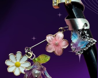 May include: A collection of decorative charms and rings. The charms include a daisy, pink flowers, a purple and pink flower, and a blue butterfly. A gold letter "M" is encrusted with rhinestones.