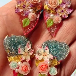 May include: Four heart-shaped resin charms featuring embedded glitter, gold leaf flakes, and miniature floral embellishments. The top two charms are pink with peach roses, while the bottom two are teal and red with coral and mint roses.