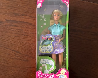 May include: A Barbie doll wearing a blue and white shirt and a purple and white floral skirt. She is holding a basket with a pink egg that says "Surprises inside egg!" The doll is part of a special edition Easter Surprise Barbie set. The box is pink and green with a picture of a chick and a bunny.