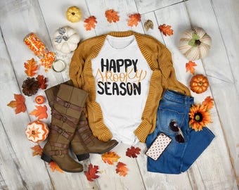 May include: A flat lay featuring a white t-shirt with "Happy Spooky Season" text, a mustard yellow cardigan, blue jeans, brown boots, and fall-themed decorations like pumpkins and leaves. Sunglasses and a phone are also included.