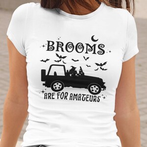 May include: White t-shirt with a black graphic featuring a Jeep with a witch and bats. The text reads "Brooms are for amateurs". A crescent moon and stars complete the Halloween design.