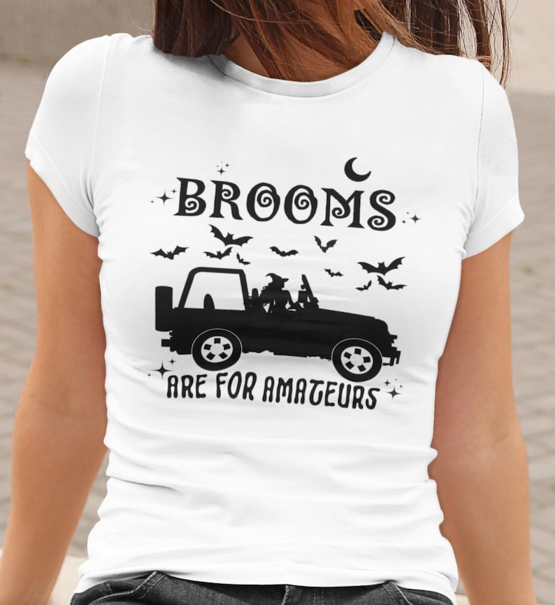 May include: White t-shirt with a black graphic featuring a Jeep with a witch and bats. The text reads "Brooms are for amateurs". A crescent moon and stars complete the Halloween design.
