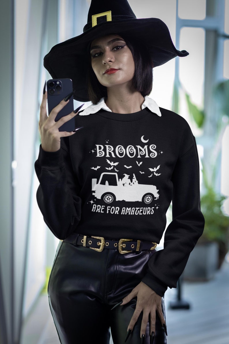 May include: Black sweatshirt with the text "Brooms Are For Amateurs" and a graphic of a truck carrying witches and bats. The model is wearing a black witch hat and black pants with a gold buckle belt. The sweatshirt has a white collar.