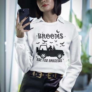 May include: White sweatshirt with the text "BROOMS ARE FOR AMATEURS" in black, featuring a jeep graphic and bats. The model is wearing a black witch hat and black pants with a gold buckle belt. A Halloween-themed apparel item.