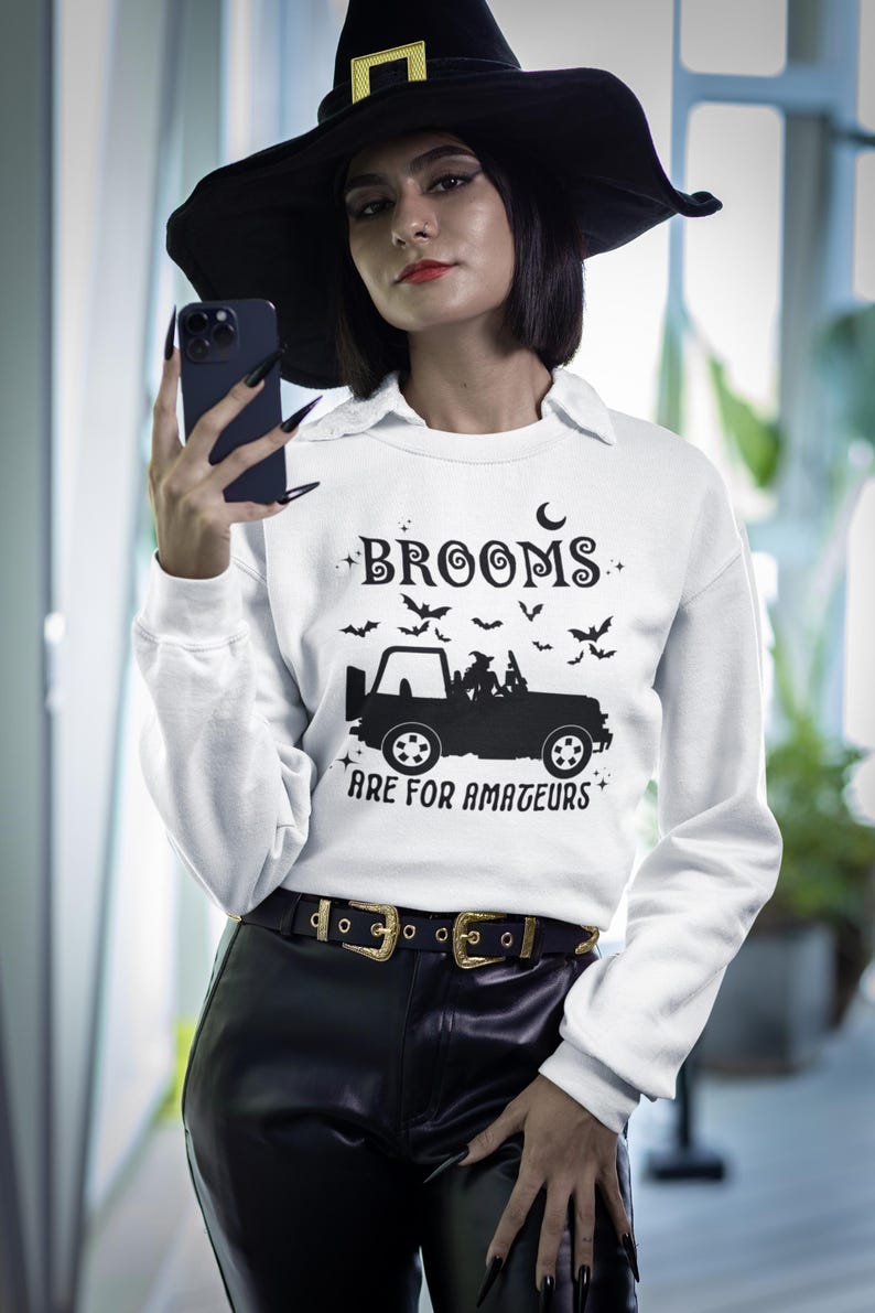 May include: White sweatshirt with the text "BROOMS ARE FOR AMATEURS" in black, featuring a jeep graphic and bats. The model is wearing a black witch hat and black pants with a gold buckle belt. A Halloween-themed apparel item.