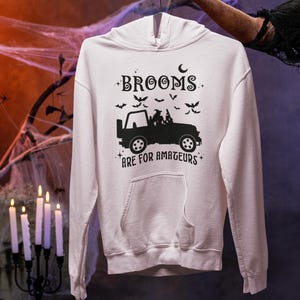 May include: A white hooded sweatshirt with a black graphic design. The design features a silhouette of a jeep with passengers, bats, and the text "BROOMS ARE FOR AMATEURS". The sweatshirt has a front pocket and a drawstring hood.