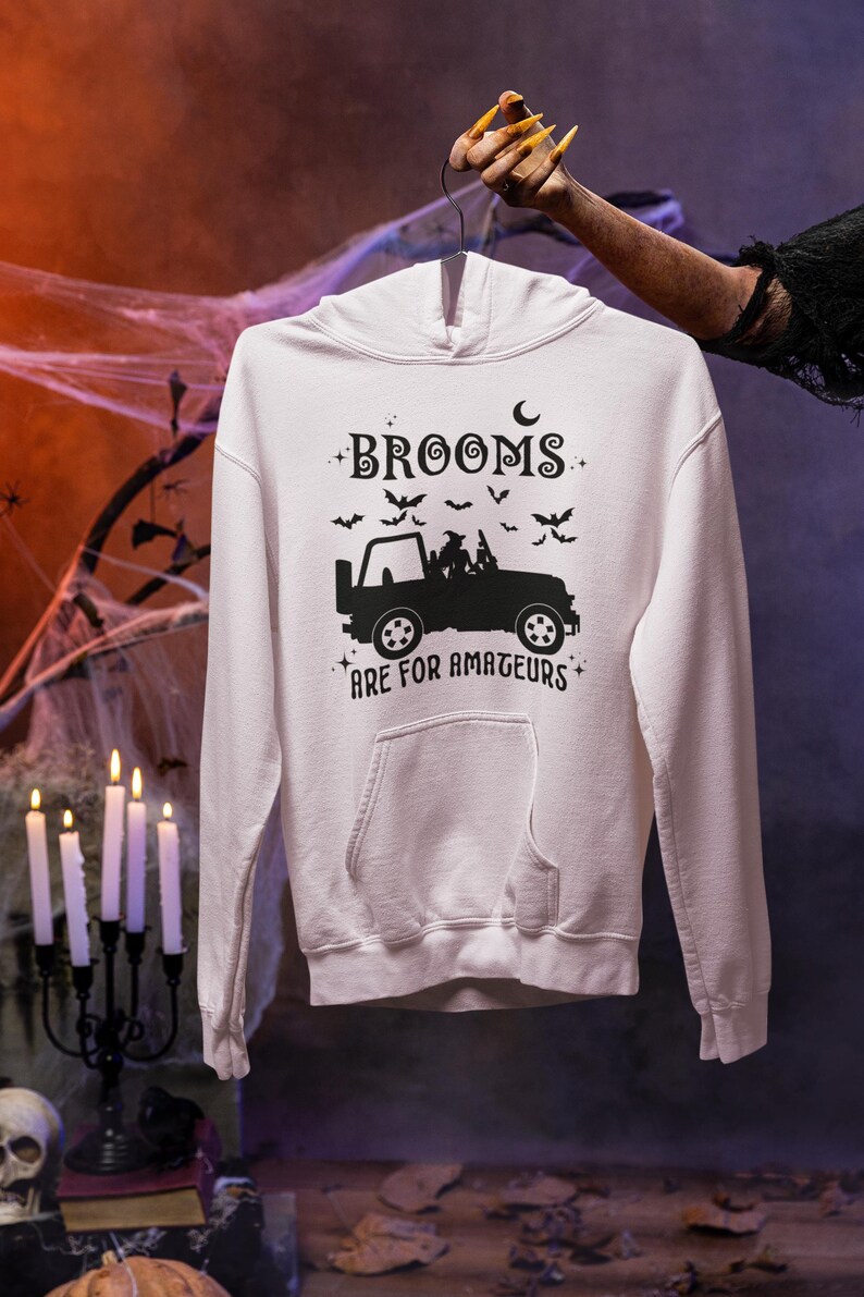 May include: A white hooded sweatshirt with a black graphic design. The design features a silhouette of a jeep with passengers, bats, and the text "BROOMS ARE FOR AMATEURS". The sweatshirt has a front pocket and a drawstring hood.