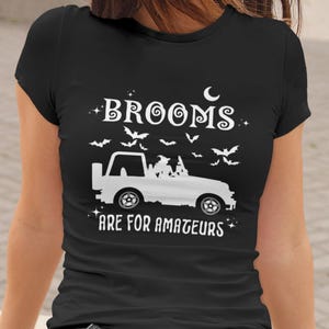 May include: Black t-shirt with a white graphic design. The design includes the text "Brooms are for amateurs" with a white jeep, bats, and a crescent moon. The shirt is short-sleeved.