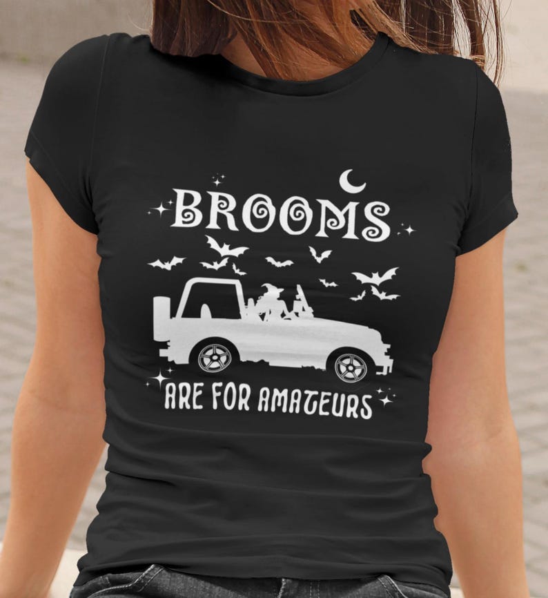 May include: Black t-shirt with a white graphic design. The design includes the text "Brooms are for amateurs" with a white jeep, bats, and a crescent moon. The shirt is short-sleeved.