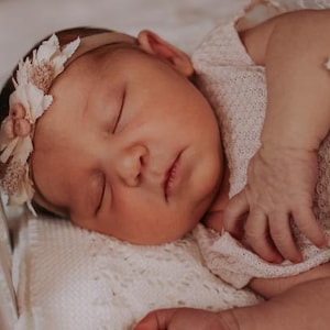 以下が含まれることがあります： 白い編み物の毛布に包まれ、眠っている新生児の女の子。白い編み物の花柄のヘッドバンドをしています。