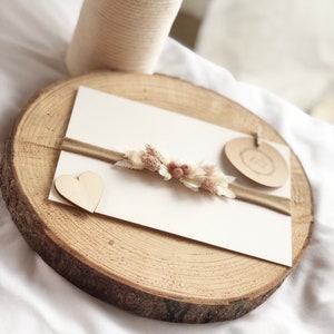 以下が含まれることがあります： 白いカードに茶色のリボンと小さな花飾り。カードの左側に小さな木製のハートが置かれている。カードは木の切り株の上に置かれている。