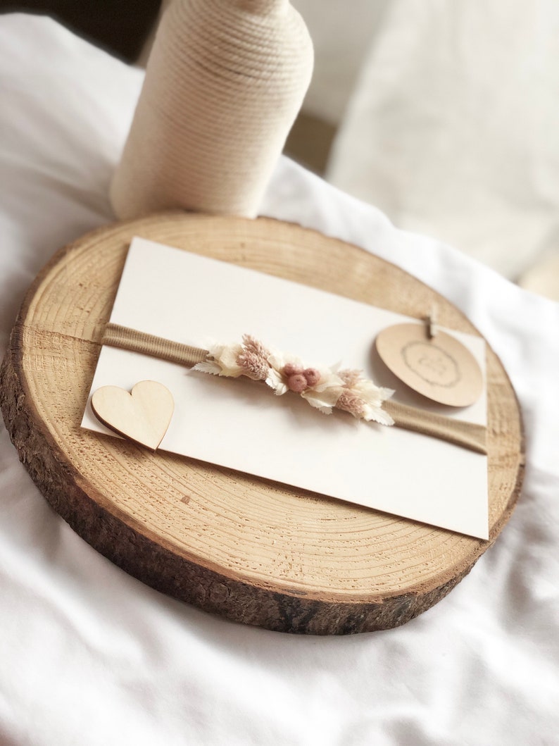 以下が含まれることがあります： 白いカードに茶色のリボンと小さな花飾り。カードの左側に小さな木製のハートが置かれている。カードは木の切り株の上に置かれている。