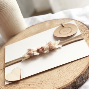以下が含まれることがあります： 中央に小さなドライフラワーとベリーの房が付いた薄い茶色のヘッドバンド。ヘッドバンドは、ハート型の木片と「Love You」という黒い文字が書かれた茶色の紙タグのある木製の表面の上に置かれています。