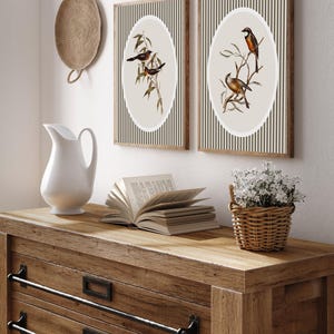 May include: Two framed bird prints with oval illustrations on a striped background. The prints are displayed on a wooden dresser with a white pitcher, an open book, and a small basket of white flowers. A woven wall hanging is also visible.