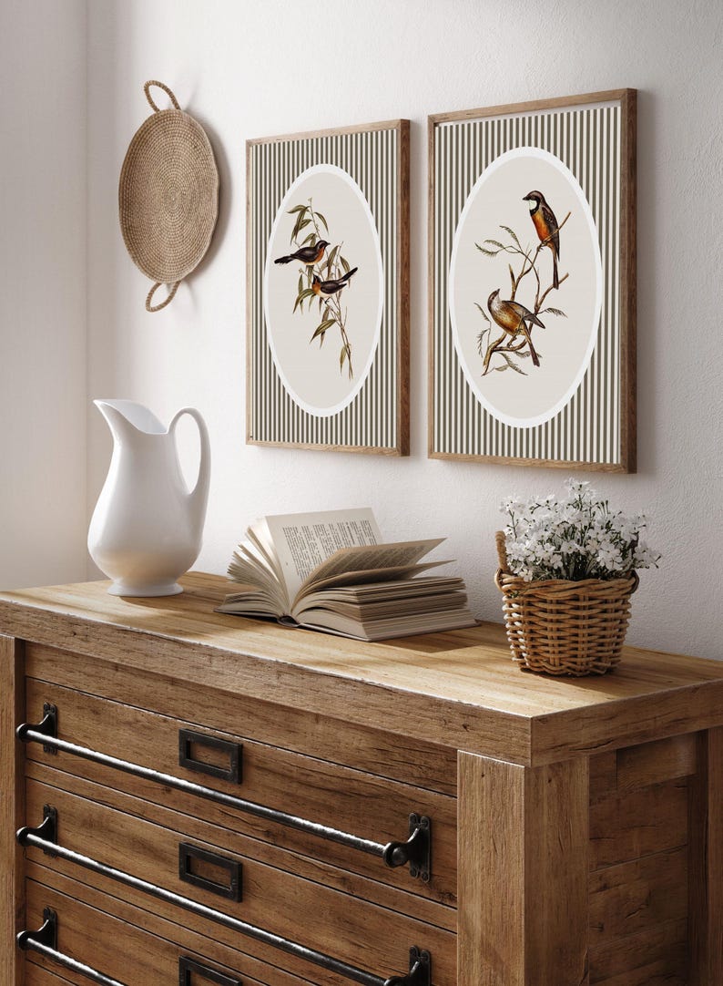 May include: Two framed bird prints with oval illustrations on a striped background. The prints are displayed on a wooden dresser with a white pitcher, an open book, and a small basket of white flowers. A woven wall hanging is also visible.