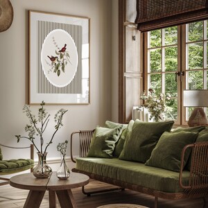 May include: A framed botanical print featuring two birds perched on a branch, set against a striped background. The artwork is displayed in a living room with a green sofa, pillows, and a wooden coffee table. A woven ottoman sits on the floor.