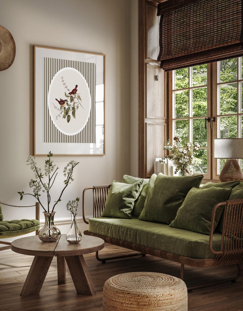 May include: A framed botanical print featuring two birds perched on a branch, set against a striped background. The artwork is displayed in a living room with a green sofa, pillows, and a wooden coffee table. A woven ottoman sits on the floor.