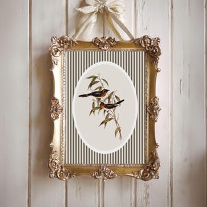 May include: A framed print featuring two birds perched on a leafy branch. The artwork is set against a striped background within an oval shape. The ornate gold frame is suspended by a cream-colored ribbon, creating a vintage aesthetic.