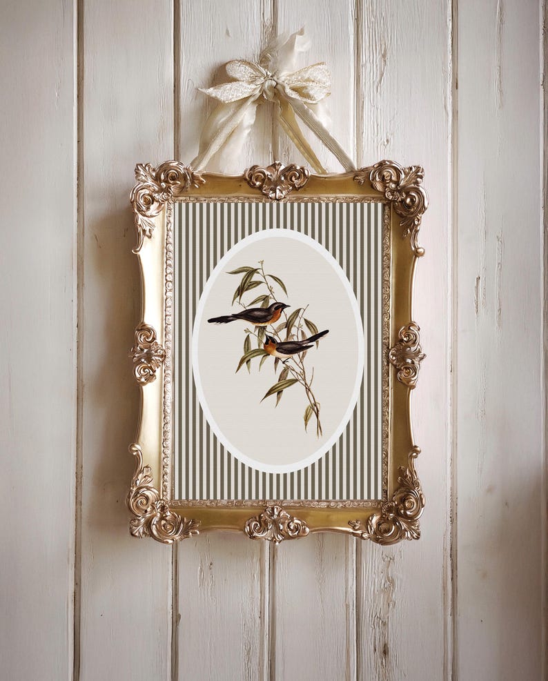 May include: A framed print featuring two birds perched on a leafy branch. The artwork is set against a striped background within an oval shape. The ornate gold frame is suspended by a cream-colored ribbon, creating a vintage aesthetic.