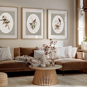 May include: Three framed wall art pieces with bird illustrations. Each features a bird perched on a branch within an oval shape, set against a striped background. The frames are a light wood color, and the overall aesthetic is neutral and natural.
