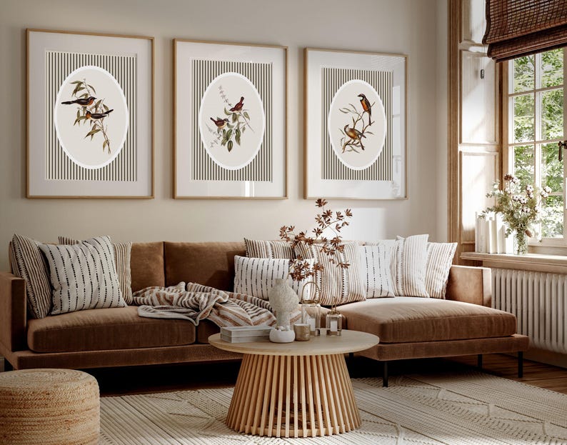 May include: Three framed wall art pieces with bird illustrations. Each features a bird perched on a branch within an oval shape, set against a striped background. The frames are a light wood color, and the overall aesthetic is neutral and natural.