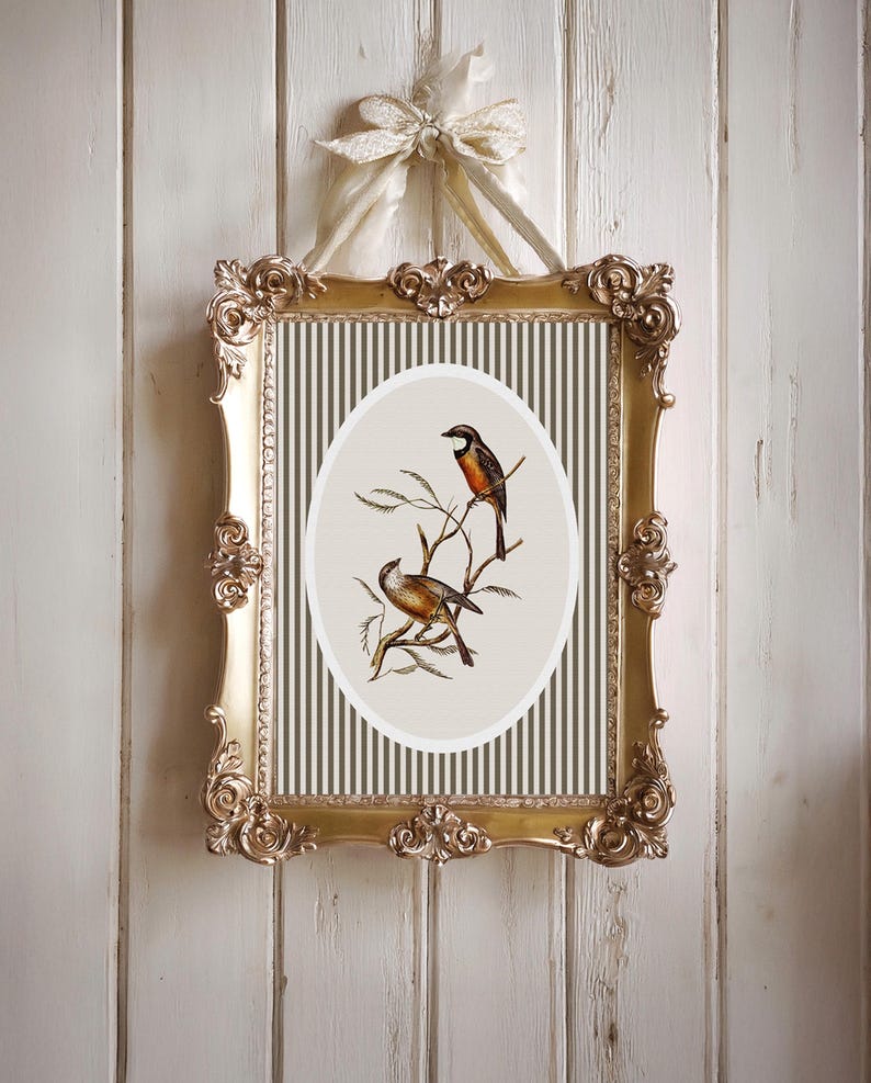 May include: Ornate gold framed artwork featuring two birds perched on branches. The artwork has a cream-colored oval center with a striped background. The frame is detailed with scrollwork and is suspended by a cream-colored ribbon.