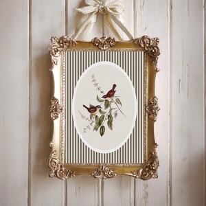 May include: Ornate gold framed artwork featuring an oval print of two birds perched on a leafy branch. The background has vertical stripes in olive green and cream. The frame is suspended by a cream-colored ribbon.