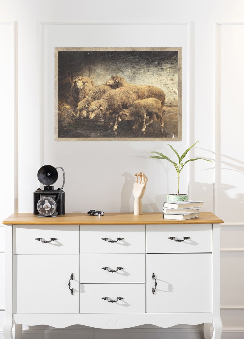 May include: A framed vintage-style print of a group of sheep in a field. The print has a distressed, textured look and is displayed on a white wall above a dresser.