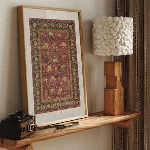 May include: A framed rectangular print featuring a deep red background with intricate gold and dark green floral and animal motifs, displayed next to a white textured pebble-style lamp and a vintage black camera on a wooden shelf.