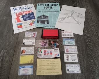 May include: A collection of items from the Back to the Future movie franchise. The items include a newspaper clipping, a flyer for a band audition, a drawing of the DeLorean time machine, a wallet, a library card, a school tardy slip, a ticket to the 'Goonies' movie, and a driver's license.