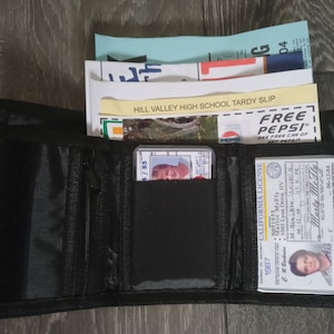 May include: A black wallet with a zipper closure, containing a driver's license, a school tardy slip, and a Pepsi coupon. The driver's license has the text "CALIFORNIA LICENSE" and the name "Marty Me Fly". The tardy slip has the text "HILL VALLEY HIGH SCHOOL TARDY SLIP". The Pepsi coupon has the text "FREE PEPSI" and "ONE FREE CAN OF ANY PEPSI".