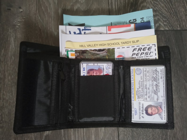 May include: A black wallet with a zipper closure, containing a driver's license, a school tardy slip, and a Pepsi coupon. The driver's license has the text "CALIFORNIA LICENSE" and the name "Marty Me Fly". The tardy slip has the text "HILL VALLEY HIGH SCHOOL TARDY SLIP". The Pepsi coupon has the text "FREE PEPSI" and "ONE FREE CAN OF ANY PEPSI".