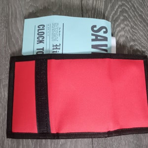 May include: Red canvas wallet with black trim and a black elastic band. The wallet is closed and has the text "Hi" and "Clock To" printed on the front.