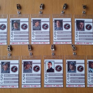 May include: A collection of twelve rectangular plastic ID badge holders with metal clips, each featuring a United Planets C-570 crew member profile, including a portrait photo, name, rank, and position, arranged in two rows on a wooden surface.