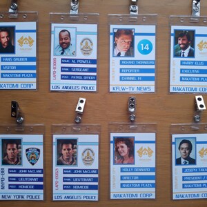 Puede incluir: Ocho porta-credenciales de plástico con rayas azules y blancas. Cada porta-credencial tiene una foto e información sobre un personaje de la película Die Hard. El texto en las credenciales incluye nombres, títulos y afiliaciones como Nakatomi Plaza, la policía de Los Ángeles y la policía de Nueva York.