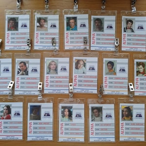 May include: A set of 15 plastic ID badges with printed images and text. The badges are labelled "ALPHA" and feature names, ranks, and roles. The badges are hung on a wall with metal clips.