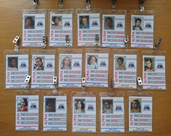 May include: A set of 15 plastic ID badges with printed images and text. The badges are labeled "ALPHA" and feature names, ranks, and roles. The badges are hung on a wall with metal clips.