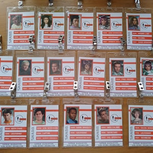 May include: A set of 15 plastic ID badges with red and white text. Each badge features a photo of a person and their name, rank, and department. The text on the badges reads "SHADO" and "Supreme Headquarters Alien Defence Organisation".