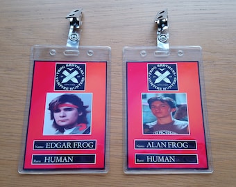 Może przedstawiać: Dwie czerwono-czarne plastikowe identyfikatory z fotografiami dwóch młodych mężczyzn. Identyfikatory zawierają tekst "Frog Brothers Vampire Hunters" oraz imiona "Edgar Frog" i "Alan Frog". Oba identyfikatory podają rasę jako "Człowiek".