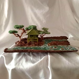 May include: A miniature diorama featuring a green pagoda-style building nestled among brown rock formations and green trees. The scene includes a small body of water with a textured, wave-like appearance. The base is a dark brown, rectangular shape.