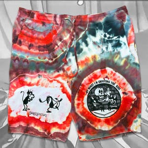 May include: Tie-dye shorts with a drawstring. The shorts display a vibrant mix of red, orange, blue, and green colours. They feature two cartoon graphics: a devil and a horse, and a skeleton with a cartoon animal. The text "AT LEAST I'M ENJOYING THE RIDE" is visible.