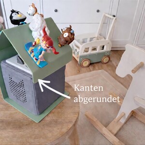 May include: A gray speaker box with a green roof, topped with toy figurines. A wooden rocking horse and a toy bus are in the background. The box has rounded edges.