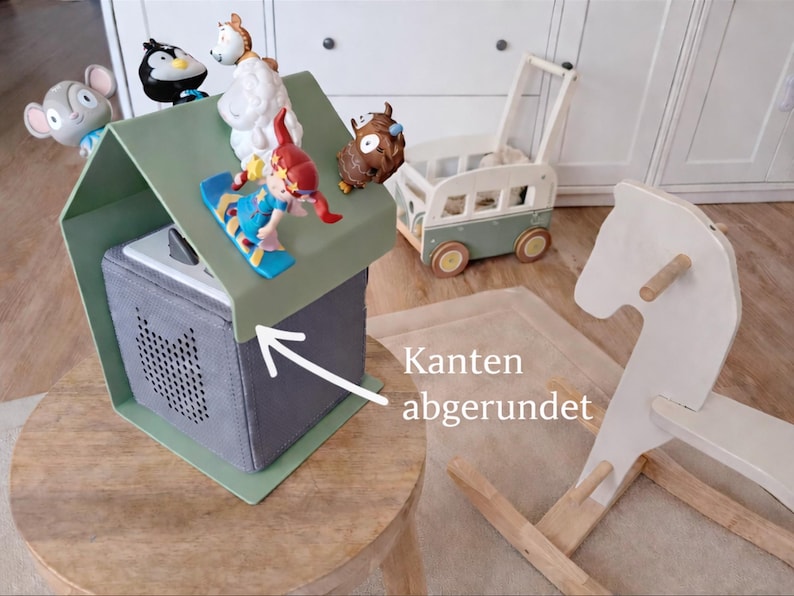 May include: A gray speaker box with a green roof, topped with toy figurines. A wooden rocking horse and a toy bus are in the background. The box has rounded edges.